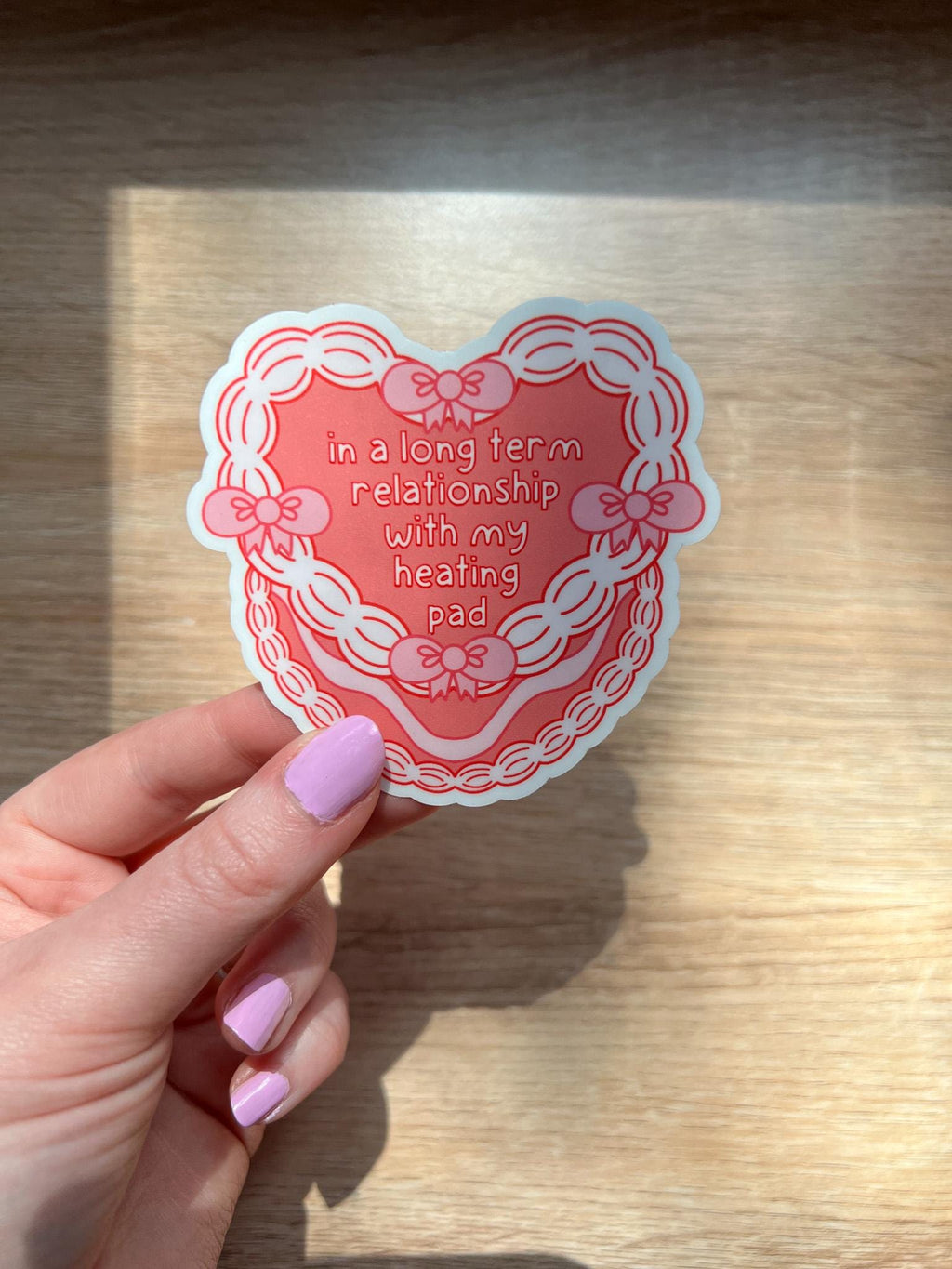 Heart cake shaped sticker. Heart cake is a bright coral color decorated with white frosting and pink bows around the front of the cake. The text outlined in dark red and filled in with white reads, "in a long term relationship with my heating pad". Sticker is held over wooden desk.