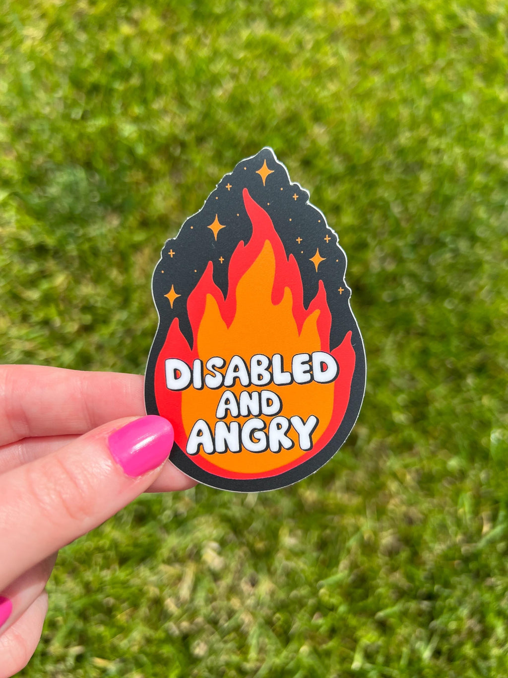 Flame shaped sticker with black background, an orange and red flame with sparkles around the top. The font in the flames center reads, "disabled and angry". The sticker is held over grass on a sunny day. 