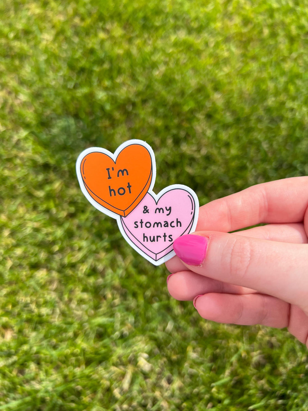 Candy heart sticker with one heart being a bright shade of orange and the other in light pink. The bright orange heart reads, "I'm hot" while the light prink heart reads, "and my stomach hurts". Sticker is held in front of grass. 