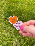 Candy heart sticker with one heart being a bright shade of orange and the other in light pink. The bright orange heart reads, "I'm hot" while the light prink heart reads, "and my stomach hurts". Sticker is held in front of grass. 
