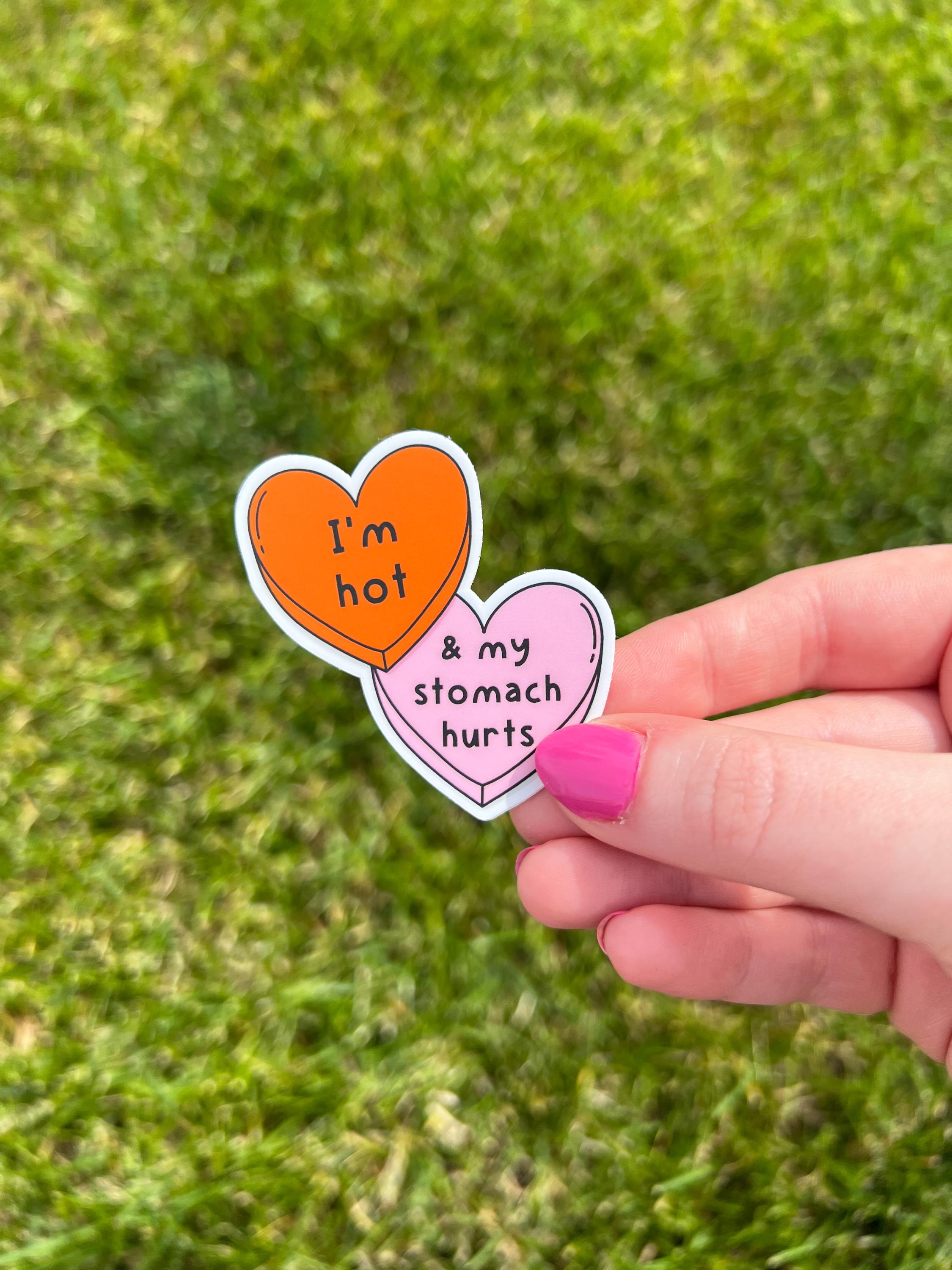 Candy heart sticker with one heart being a bright shade of orange and the other in light pink. The bright orange heart reads, "I'm hot" while the light prink heart reads, "and my stomach hurts". Sticker is held in front of grass. 