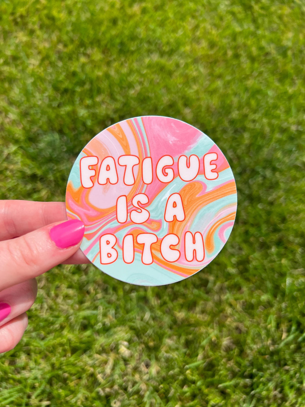 Round sticker in tie dye colors of pink, yellow orange, light blue, and white with large bubble letters outlined in burnt orange and filled in with a cream color reading, "fatigue is a bitch". Sticker is being held over grass on a sunny day. 