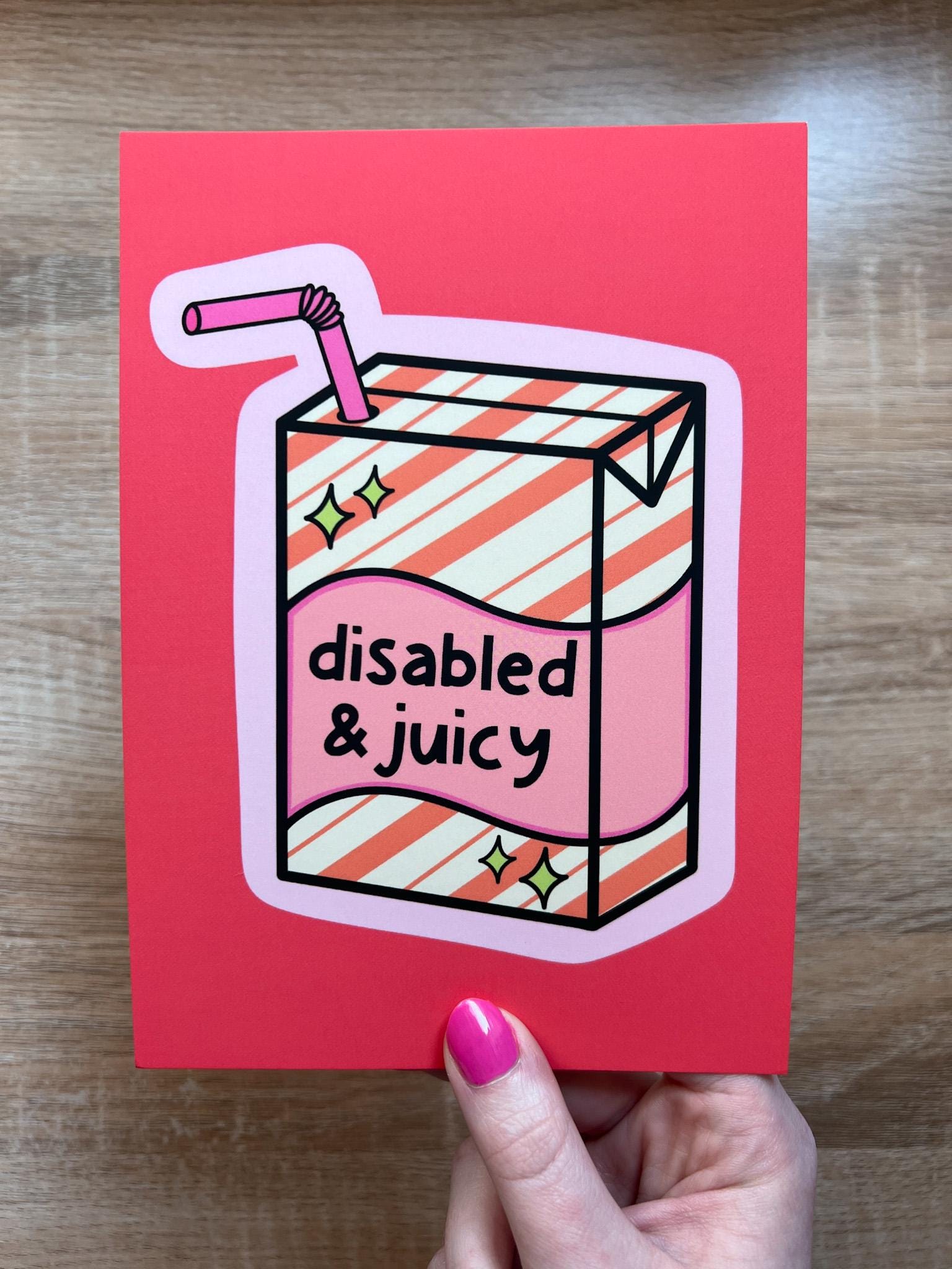 Art print with bright coral background with juice box in the center outlined in light pink. Juice box is cream with orange diagonal stripes and pink banner in the middle with yellow sparkles in either side. A pink straw is coming from the top. The black font on the juice box reads, "disabled and juicy". Print is being held over wooden desk.