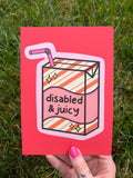 Art print with bright coral background with juice box in the center outlined in light pink. Juice box is cream with orange diagonal stripes and pink banner in the middle with yellow sparkles in either side. A pink straw is coming from the top. The black font on the juice box reads, "disabled and juicy". Print is being held over grass on a sunny day. 