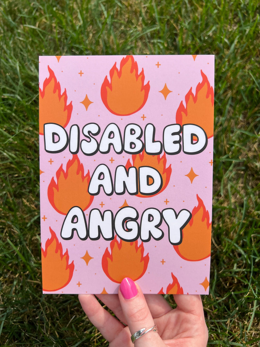 Art print with light pink background and flames in red and orange with sparkles around them with bubble letters outlined in black and filled in with white that read, "disabled and angry". The print is held over grass on a sunny day. 