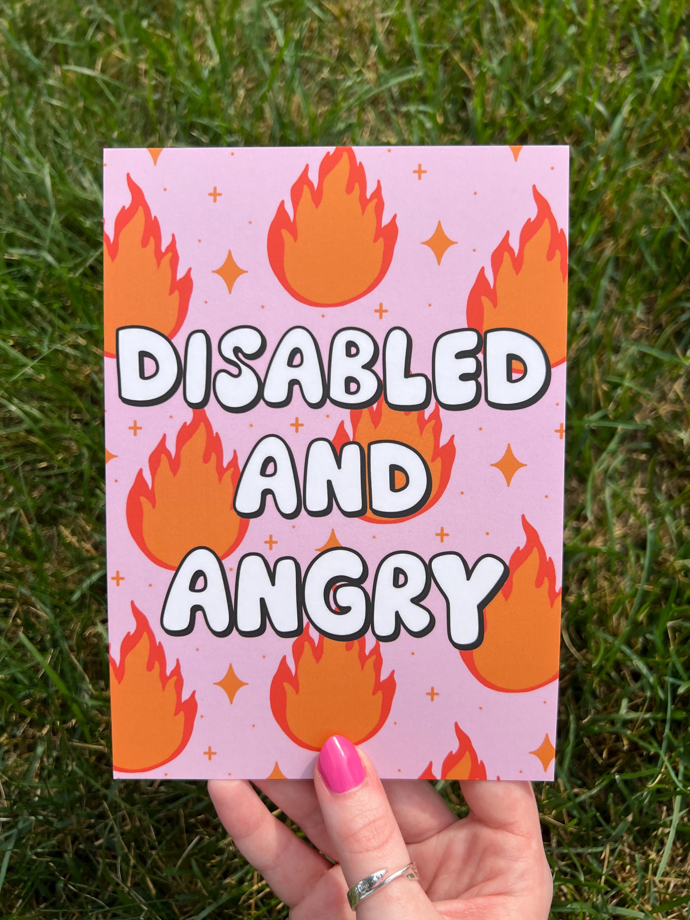 Art print with light pink background and flames in red and orange with sparkles around them with bubble letters outlined in black and filled in with white that read, "disabled and angry". The print is held over grass on a sunny day. 