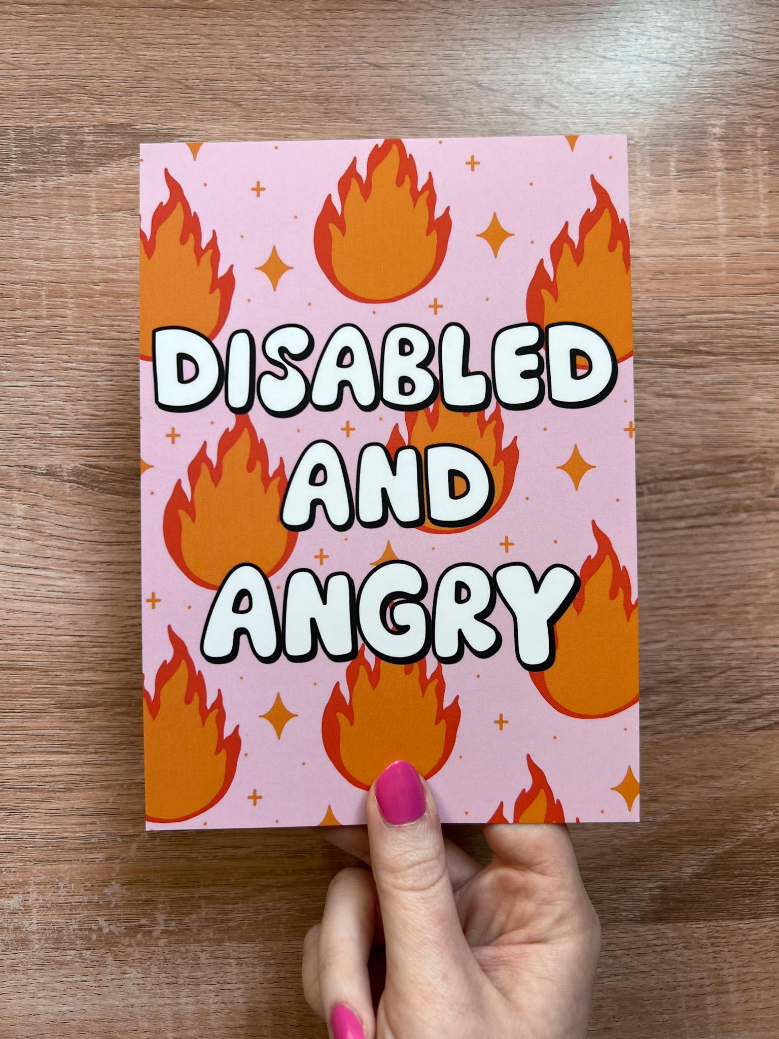 Art print with light pink background and flames in red and orange with sparkles around them with bubble letters outlined in black and filled in with white that read, "disabled and angry". The print is held over wooden desk.
