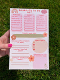 Disability To Do List Notepad with light colored sheets. Sections include: pink cloud to write the date down in, three pink boxes with red font reading "morning, noon, and night" with a checklist underneath them reading "took meds, drank water, ate something, brushed teeth, washed face" and a couple of empty spaces below to fill out yourself. There's a place to write down medical appointments to make, contact doctor about, and contact insurance about. There's a place to track symptoms. Pictured in grass.