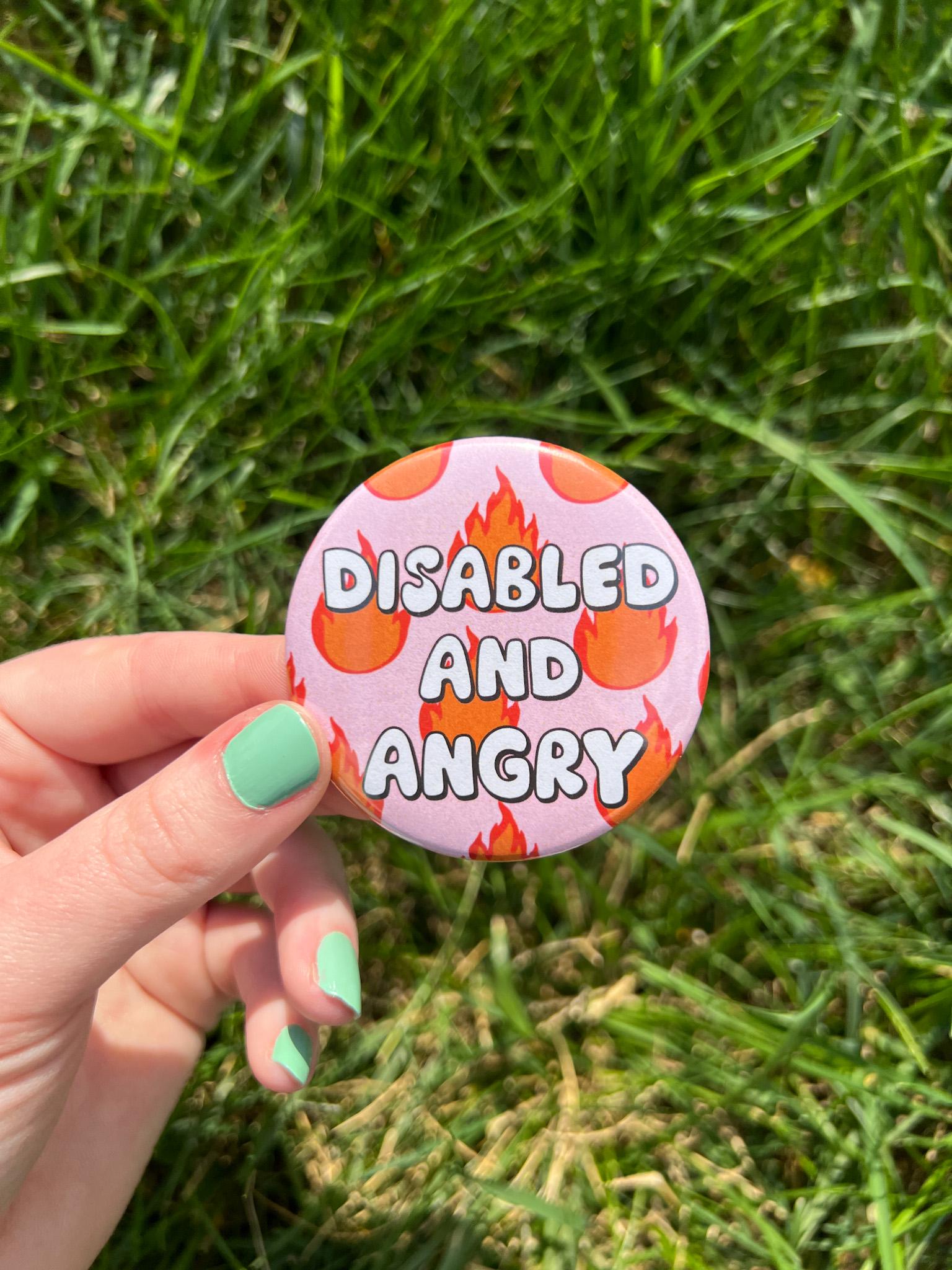 Button badge with light pink background, with red and yellow flames around it. The text in the center reads, "disabled and angry". The button is being held over grass. 