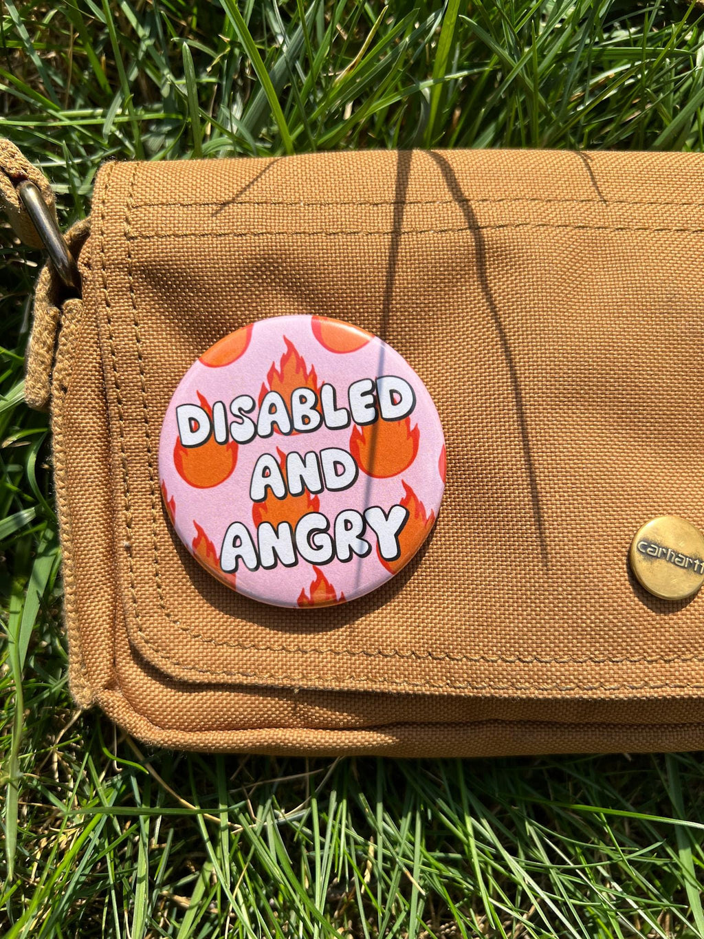 Button badge with light pink background, with red and yellow flames around it. The text in the center reads, "disabled and angry". The button is pinned to a brown bag, close up. 