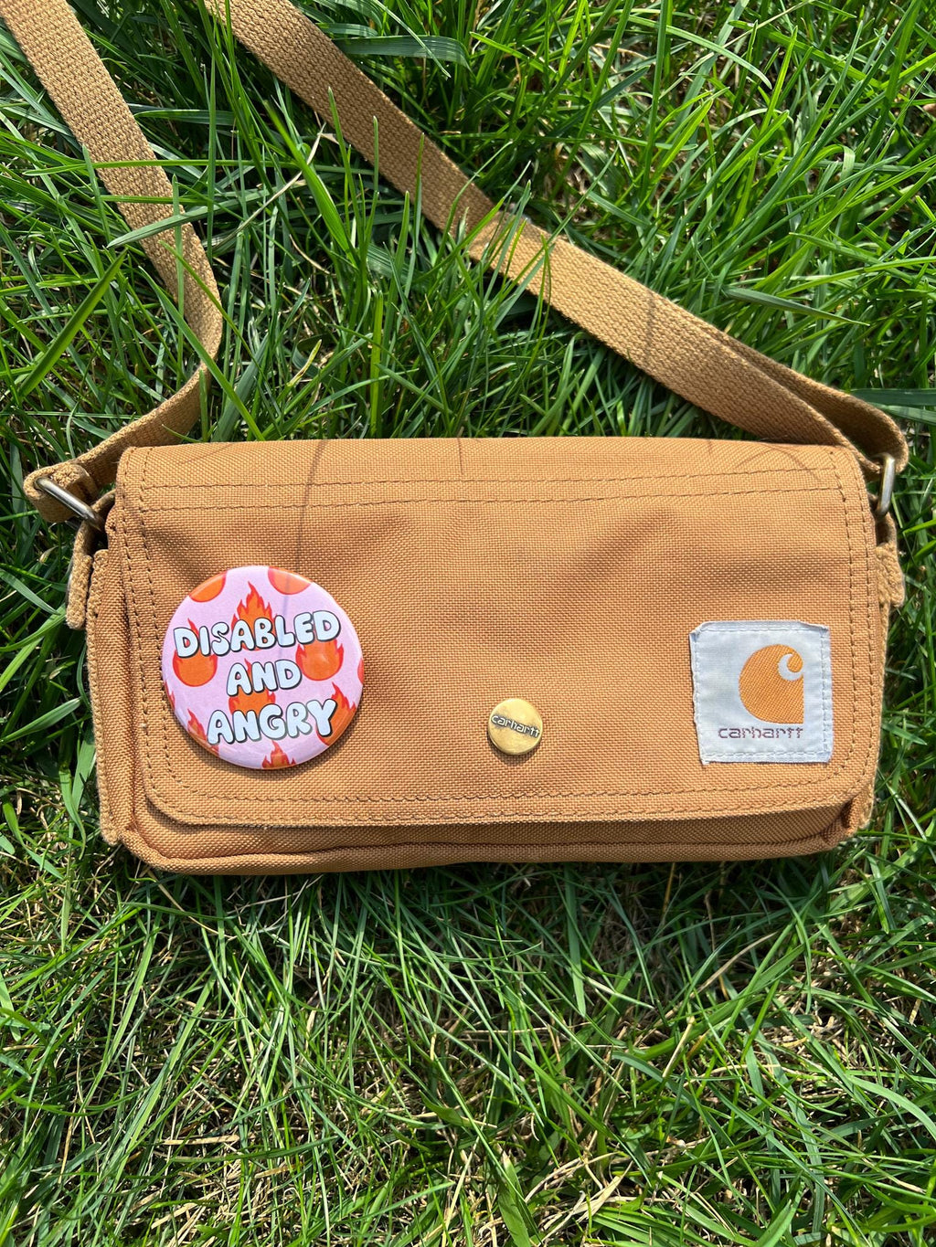 Button badge with light pink background, with red and yellow flames around it. The text in the center reads, "disabled and angry". The button is pinned to a brown bag, further away.