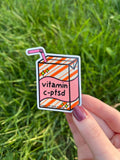 Juice box sticker, Juice box is cream with orange diagonal stripes and pink banner in the middle with yellow sparkles in either side. A pink straw is coming from the top. The black font on the juice box reads, "vitamin c-ptsd". Sticker is being held over grass on a sunny day.