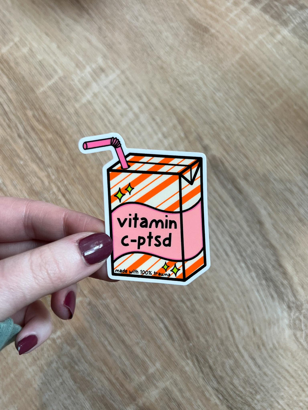 Juice box sticker, Juice box is cream with orange diagonal stripes and pink banner in the middle with yellow sparkles in either side. A pink straw is coming from the top. The black font on the juice box reads, "vitamin c-ptsd". Sticker is being held over wooden desk.