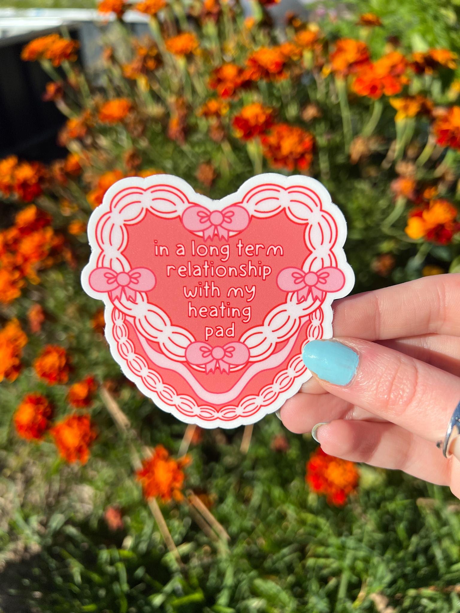 Heart cake shaped sticker. Heart cake is a bright coral color decorated with white frosting and pink bows around the front of the cake. The text outlined in dark red and filled in with white reads, "in a long term relationship with my heating pad". Sticker is held in front of orange flowers. 