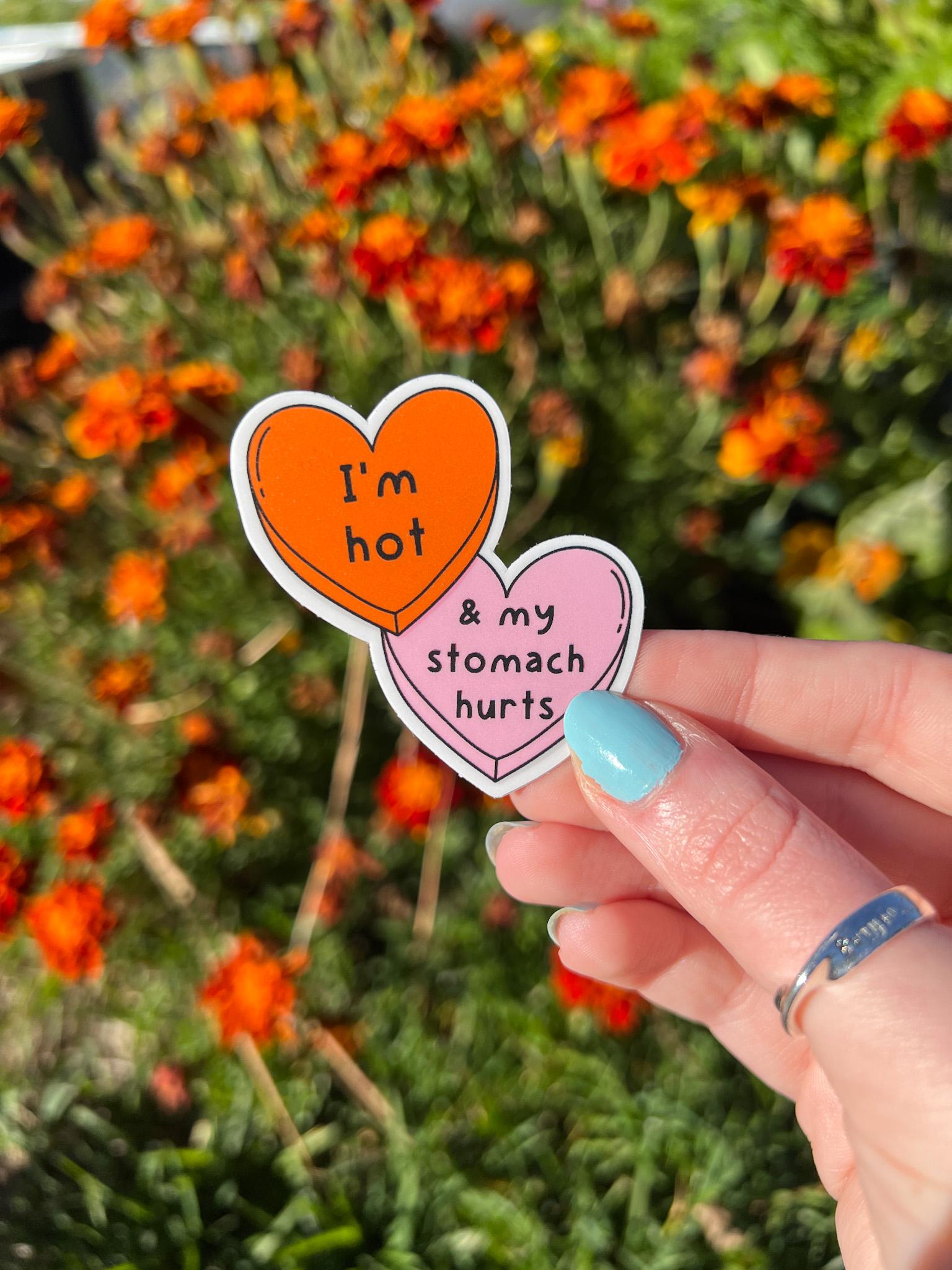 Candy heart sticker with one heart being a bright shade of orange and the other in light pink. The bright orange heart reads, "I'm hot" while the light prink heart reads, "and my stomach hurts". Sticker is held in front of orange flowers. 