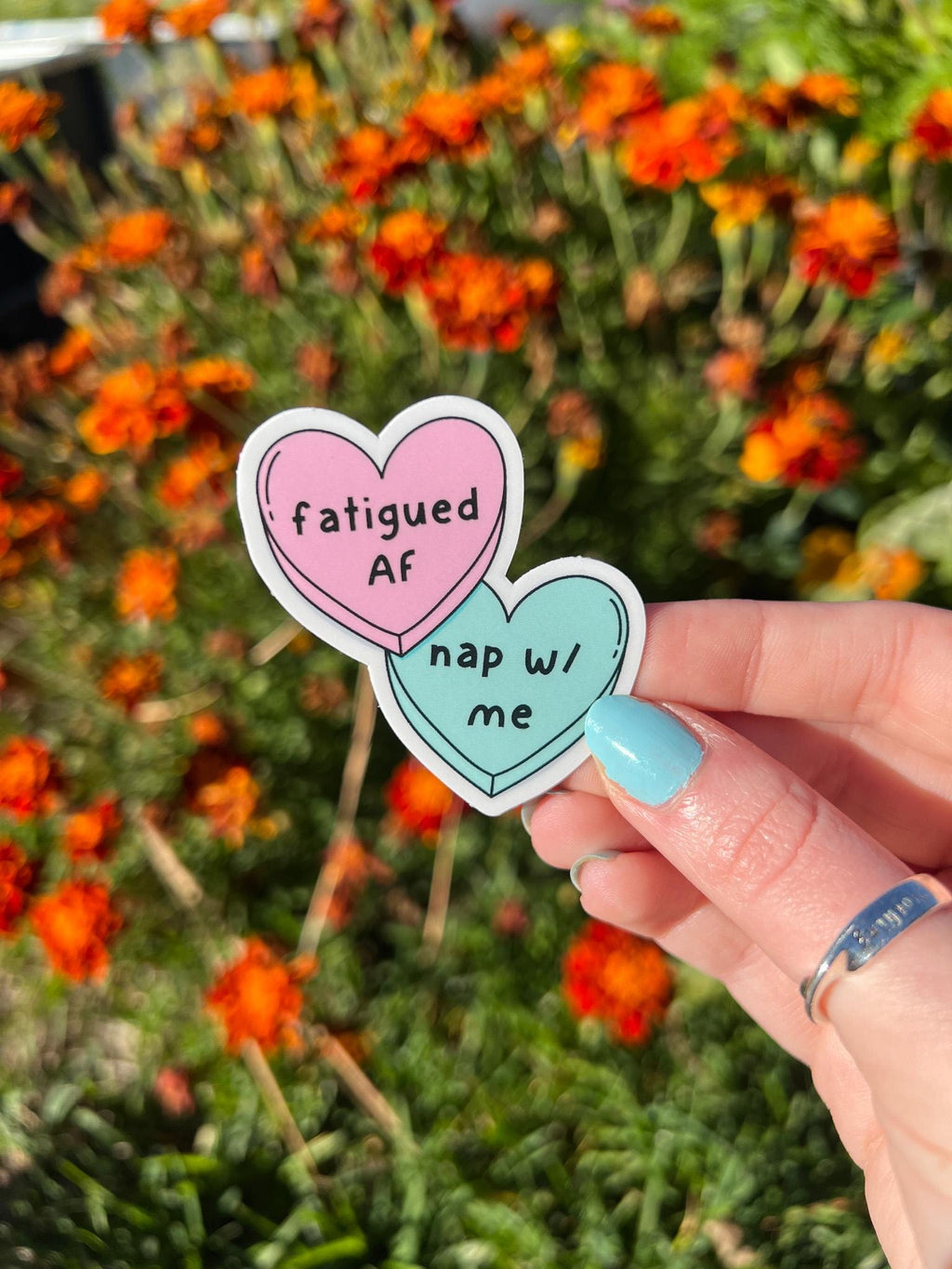 Candy heart sticker, one heart is in light pink and the other is in light blue. The font on the pink one reads, "fatigued AF" while the blue heart reads, "nap with me." Sticker is being held in front of orange flowers on a sunny day. 