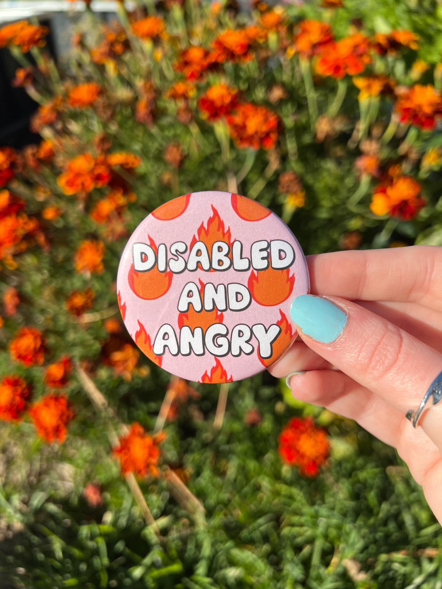 Button badge with light pink background, with red and yellow flames around it. The text in the center reads, "disabled and angry". The button is being held in front of orange flowers. 