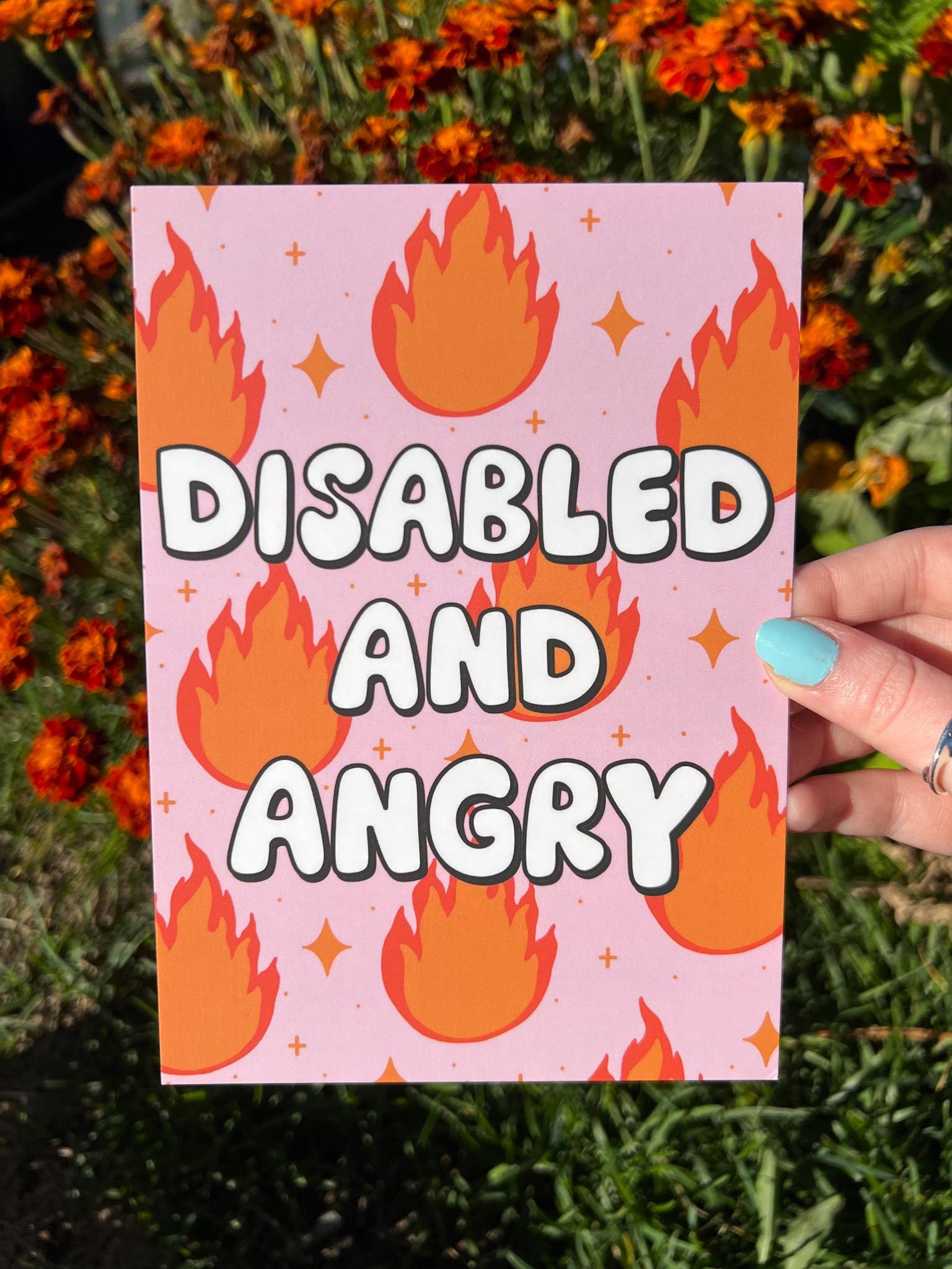 Art print with light pink background and flames in red and orange with sparkles around them with bubble letters outlined in black and filled in with white that read, "disabled and angry". The print is held in front of orange flowers on a sunny day.