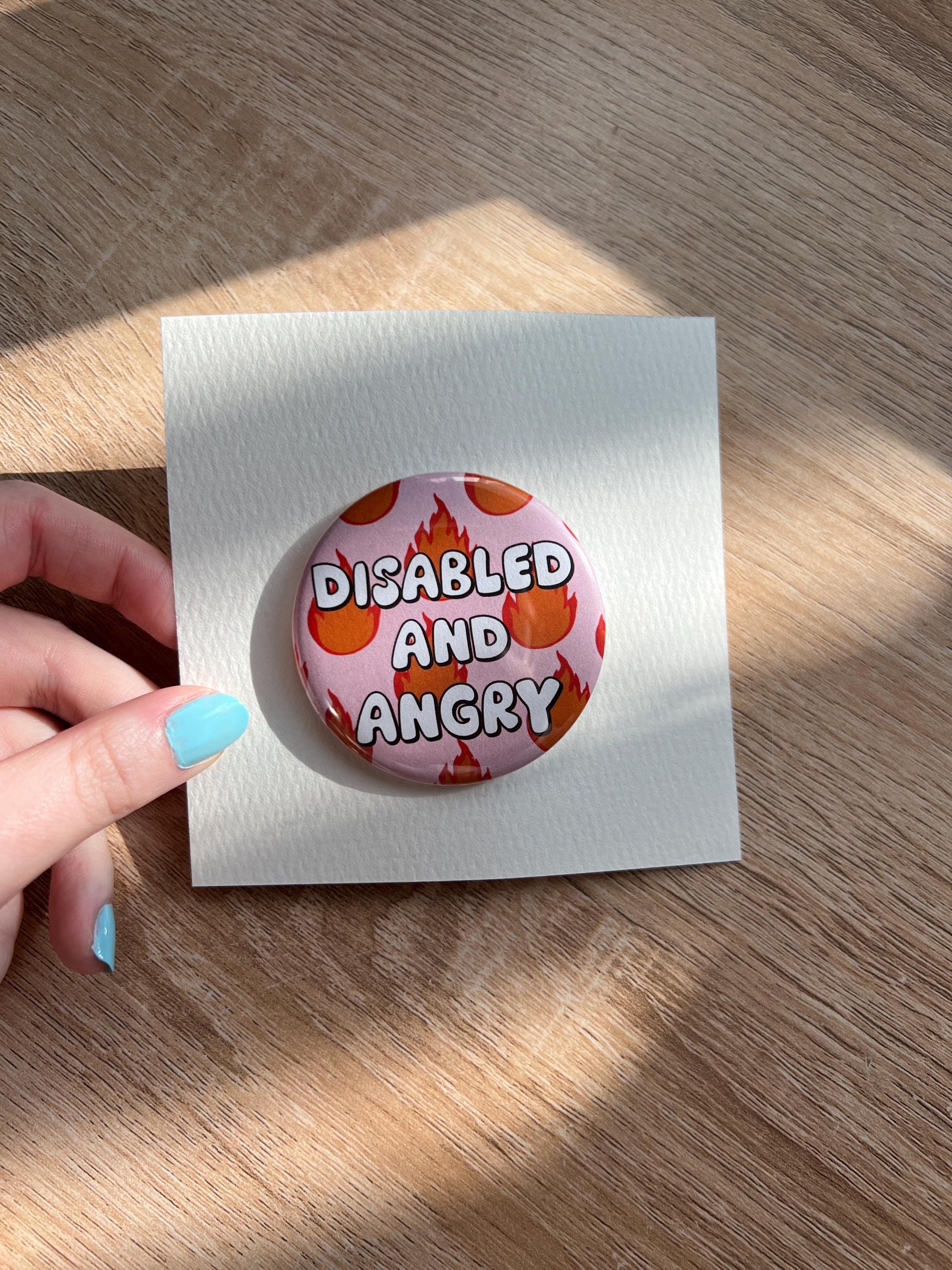 Button badge with light pink background, with red and yellow flames around it. The text in the center reads, "disabled and angry". The button is pinned to a white backing and photographed in front of wood desk. 