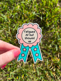 Award ribbon sticker in a light pink shade and bright turquoise with the center being cream colored. The black font reads, "fatigued AF but showed up". The sticker is being held over grass on a sunny day. 