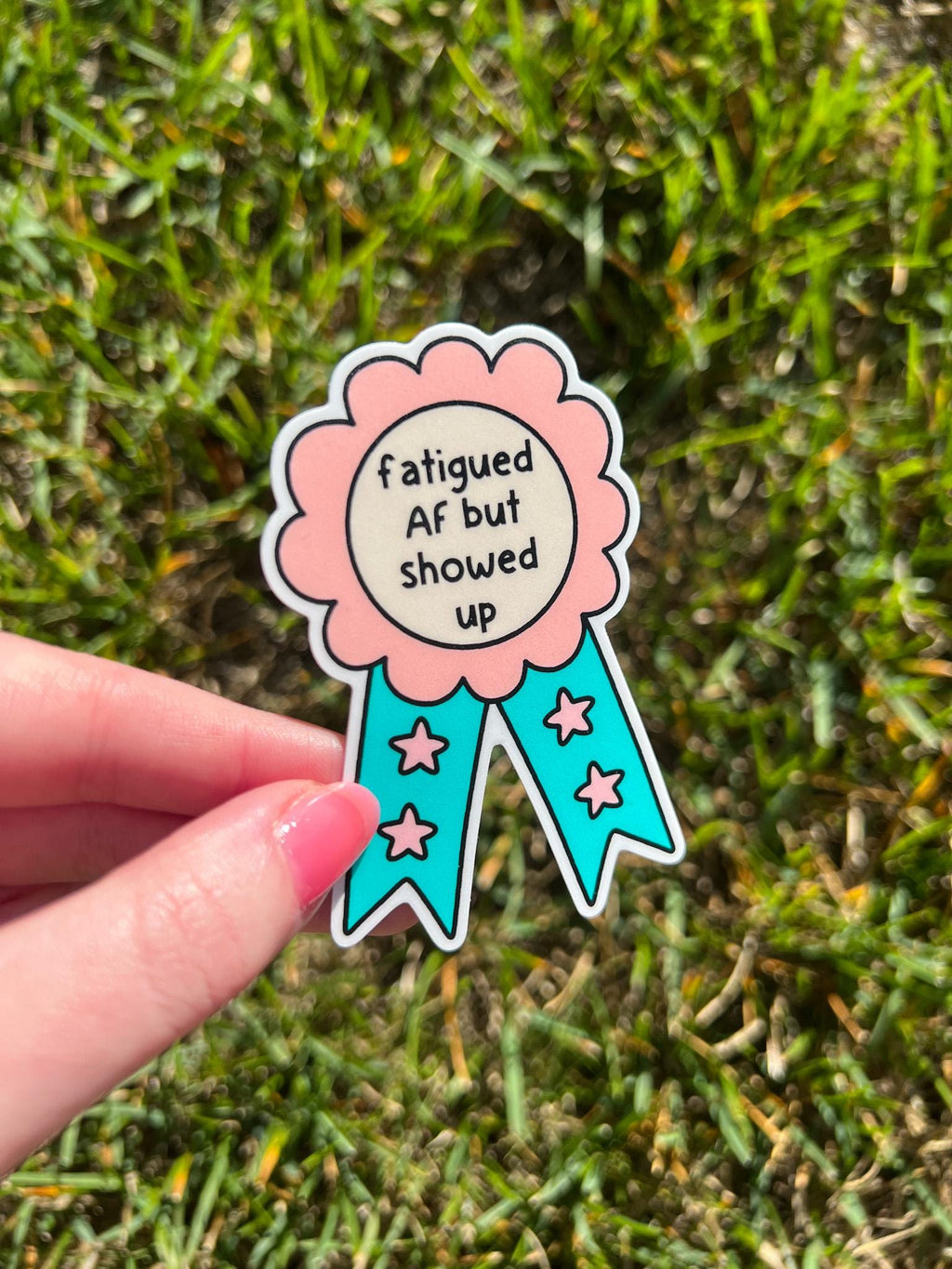 Award ribbon sticker in a light pink shade and bright turquoise with the center being cream colored. The black font reads, "fatigued AF but showed up". The sticker is being held over grass tilted to the side on a sunny day. 
