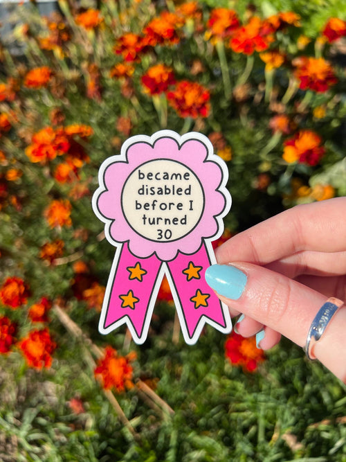 Award ribbon sticker in a light pink and dark pink shades, the middle is cream colored with the black font reading, "became disabled before I turned 30". Sticker is being held in front of orange flowers. 