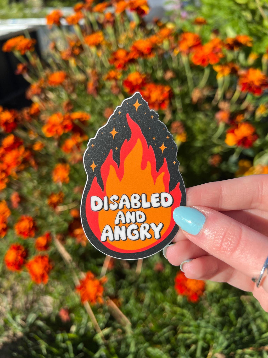 Flame shaped sticker with black background, an orange and red flame with sparkles around the top. The font in the flames center reads, "disabled and angry". The sticker is held in front of orange flowers on a sunny day. 