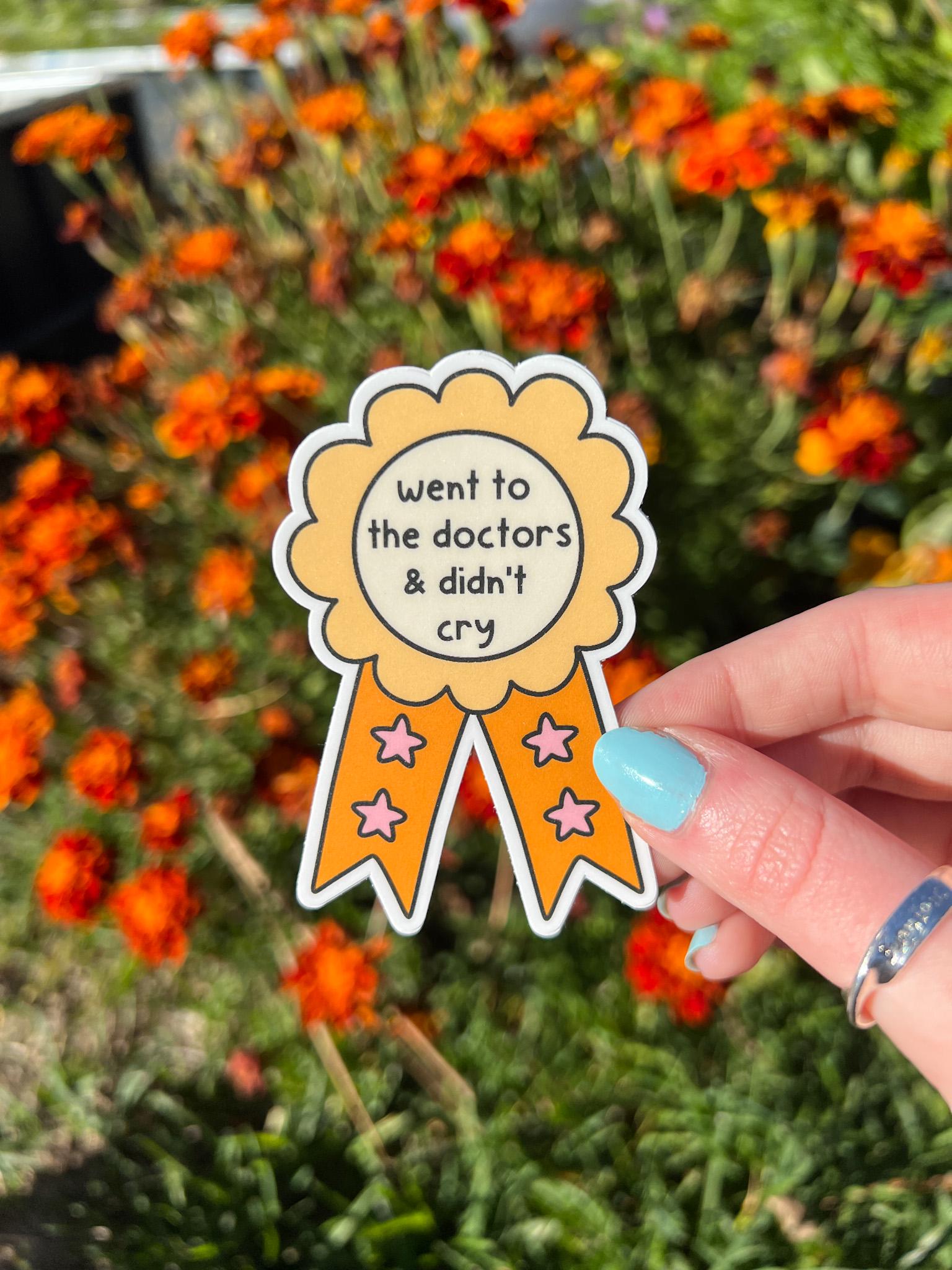Award ribbon sticker in a light shade of yellow and a bright orange with cream in the center. The black font in the center reads, "went to the doctor's and didn't cry". Sticker is held in front of orange flowers on a sunny day. 
