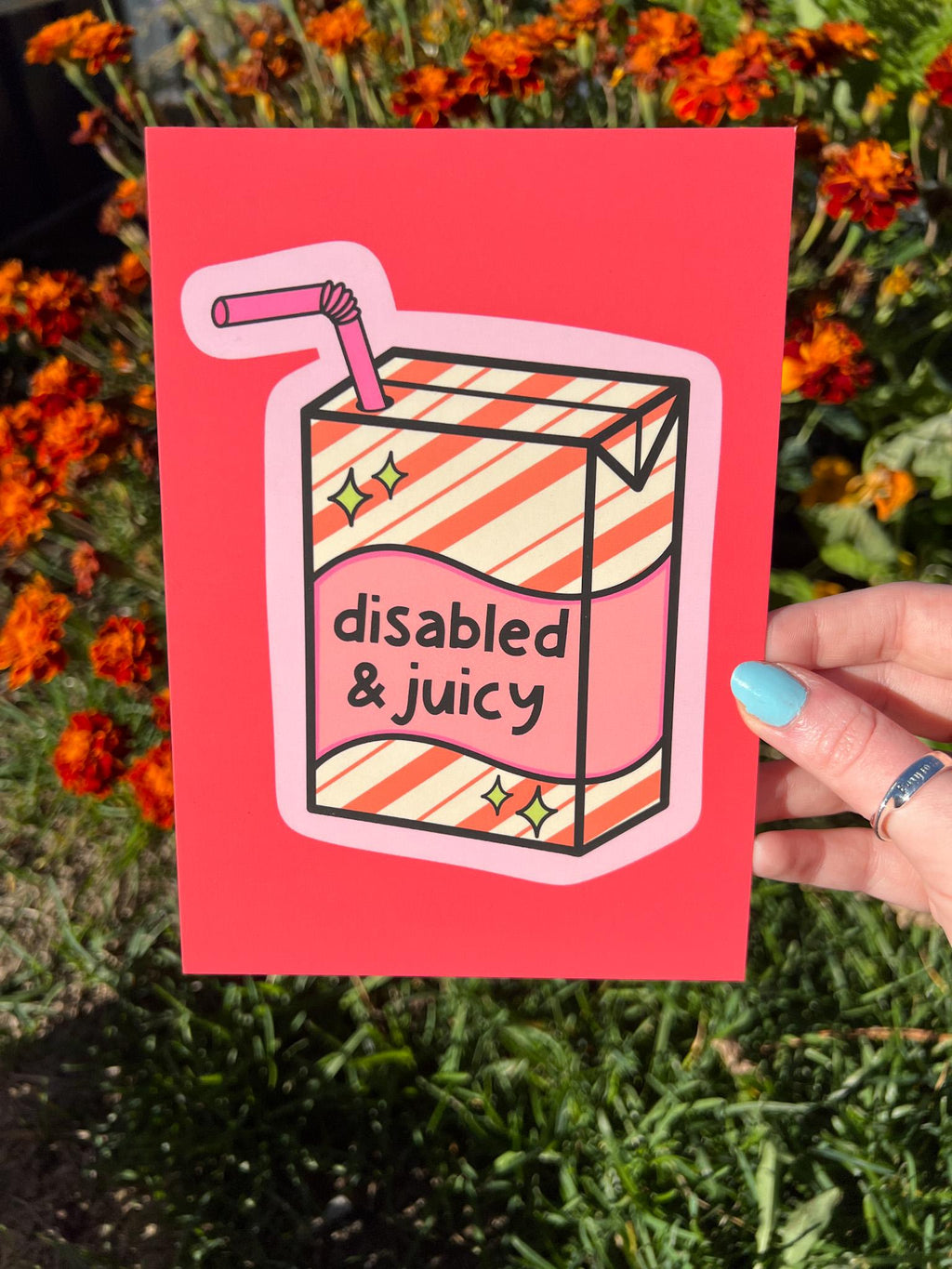 Art print with bright coral background with juice box in the center outlined in light pink. Juice box is cream with orange diagonal stripes and pink banner in the middle with yellow sparkles in either side. A pink straw is coming from the top. The black font on the juice box reads, "disabled and juicy". Print is being held in front of orange flowers on a sunny day. 
