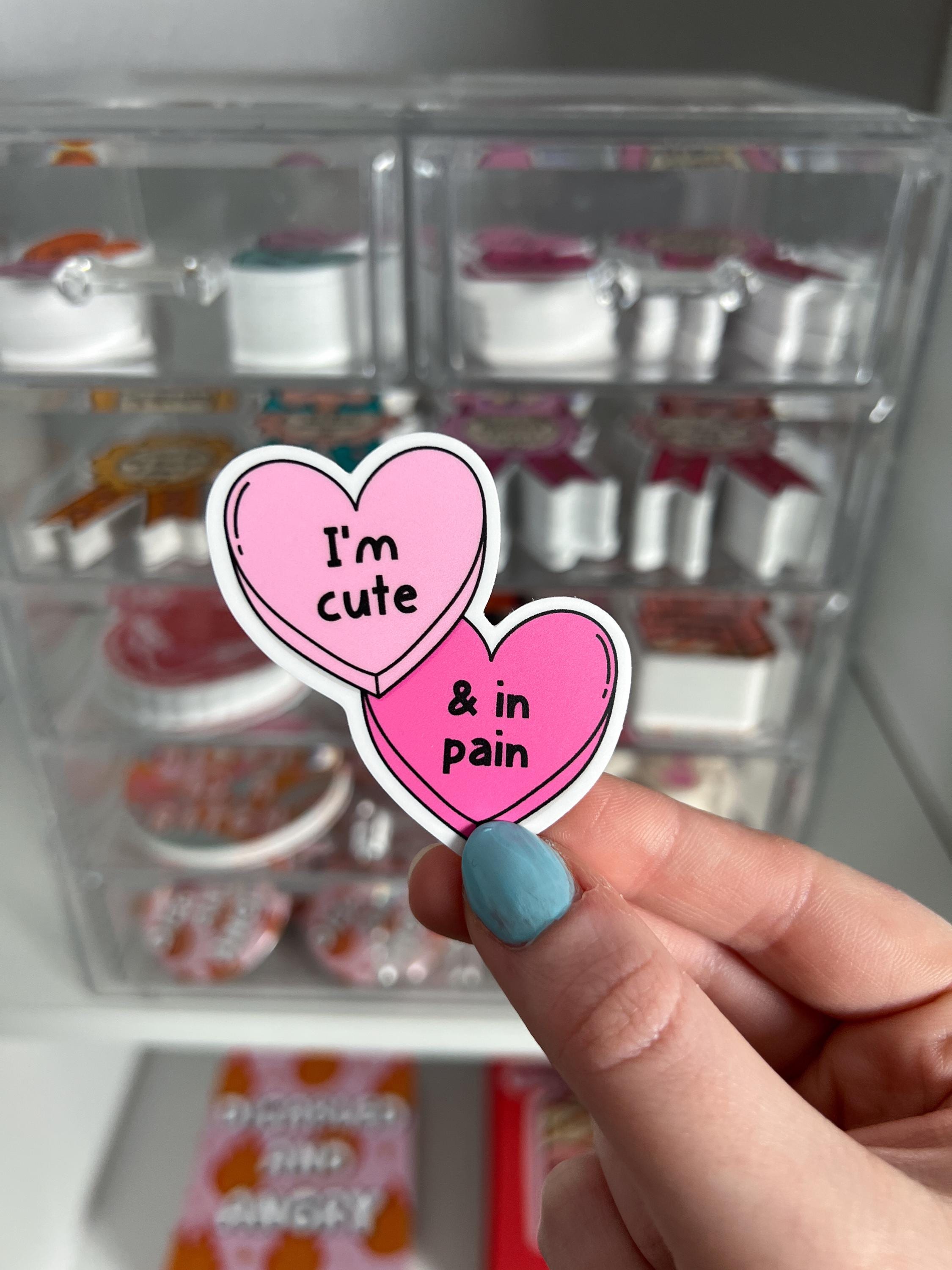 Candy hearts sticker. One heart in light pink and the other in a bright hot pink. The light pink heart reads, "I'm cute" while the hot pink reads, "and in pain". Sticker is held in front of product display. 
