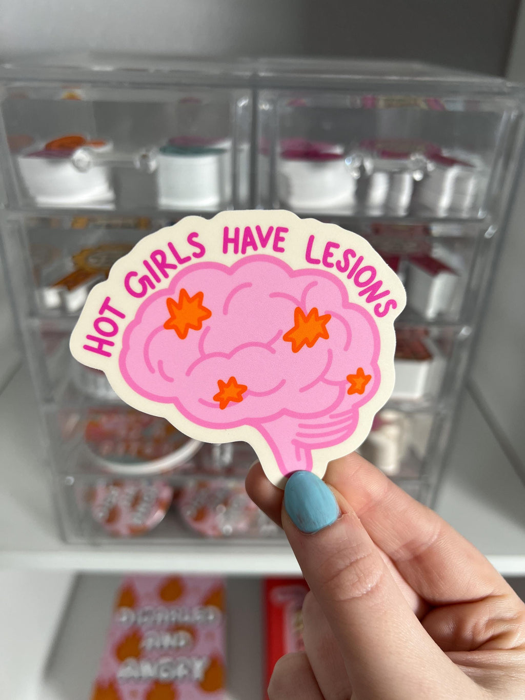 Brain shaped sticker with brain outlined in a darker bright pink and colored in with a light shade of bright pink. The brain has lesions on it that are orange. The text over the top of the brain in a hot pink reads, hot girls have lesions" with a cream background. The sticker is held in front of product display. 