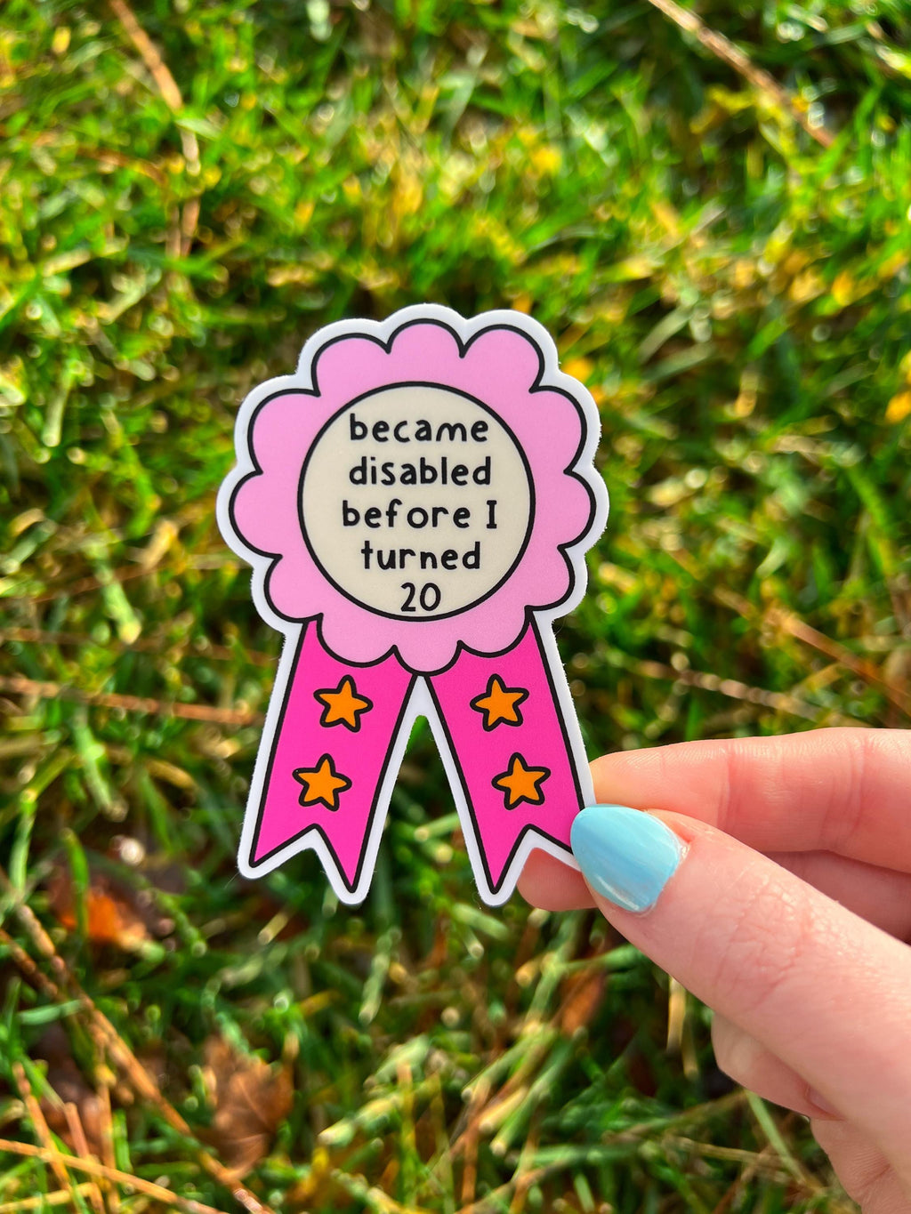 Award ribbon sticker in a light pink and dark pink shades, the middle is cream colored with the black font reading, "became disabled before I turned 20". Sticker is being held over grass. 