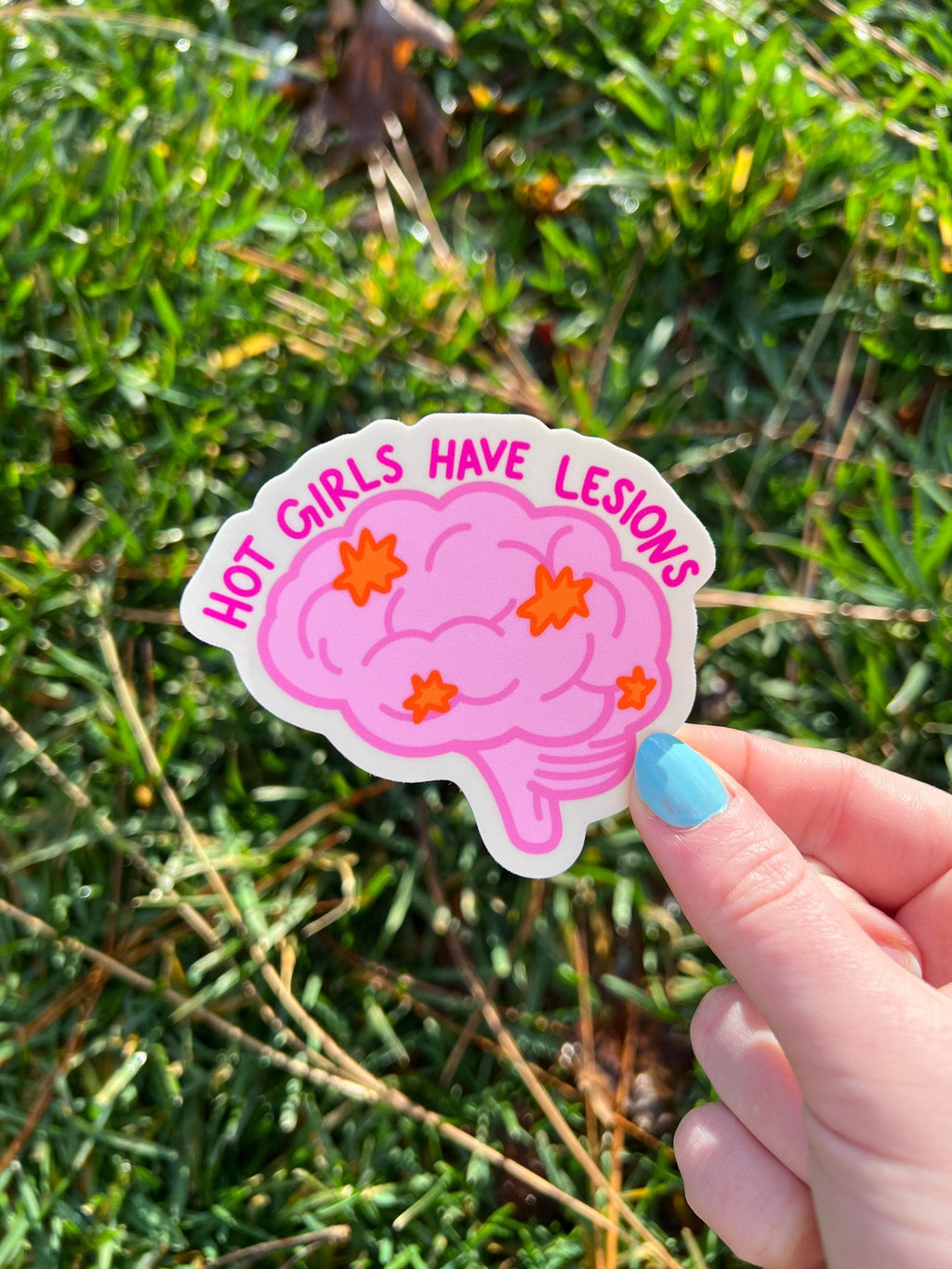 Brain shaped sticker with brain outlined in a darker bright pink and colored in with a light shade of bright pink. The brain has lesions on it that are orange. The text over the top of the brain in a hot pink reads, hot girls have lesions" with a cream background. The sticker is held over grass. 