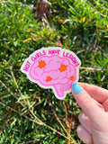 Brain shaped sticker with brain outlined in a darker bright pink and colored in with a light shade of bright pink. The brain has lesions on it that are orange. The text over the top of the brain in a hot pink reads, hot girls have lesions" with a cream background. The sticker is held over grass. 