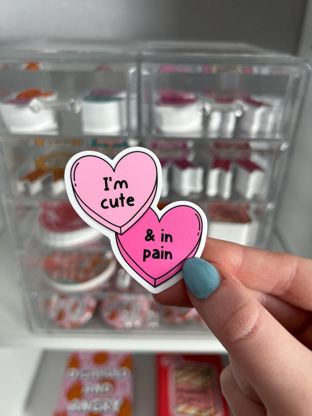 Candy hearts sticker. One heart in light pink and the other in a bright hot pink. The light pink heart reads, "I'm cute" while the hot pink reads, "and in pain". Sticker is held in front of product display. 