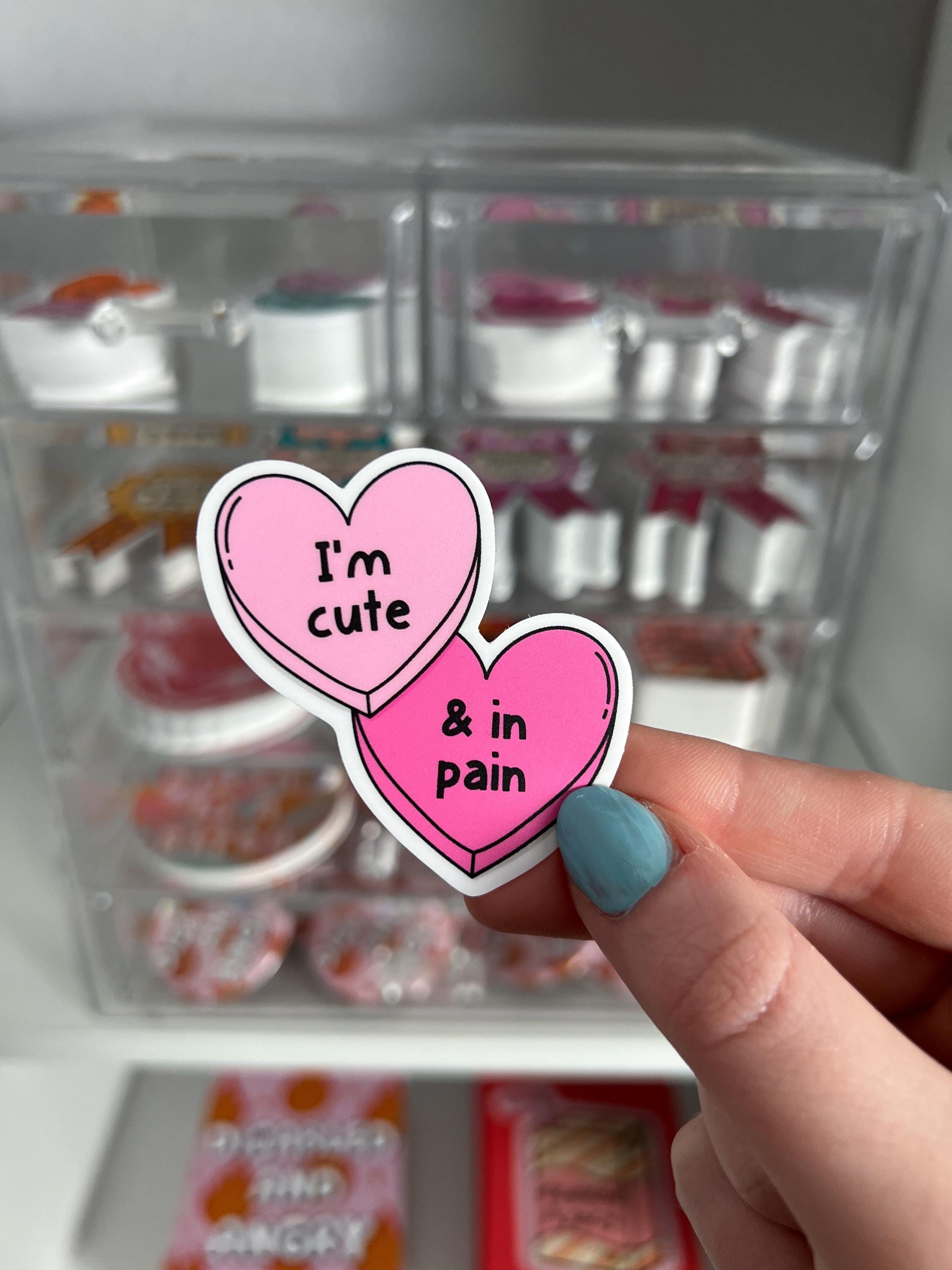 Candy hearts sticker. One heart in light pink and the other in a bright hot pink. The light pink heart reads, "I'm cute" while the hot pink reads, "and in pain". Sticker is held in front of product display. 