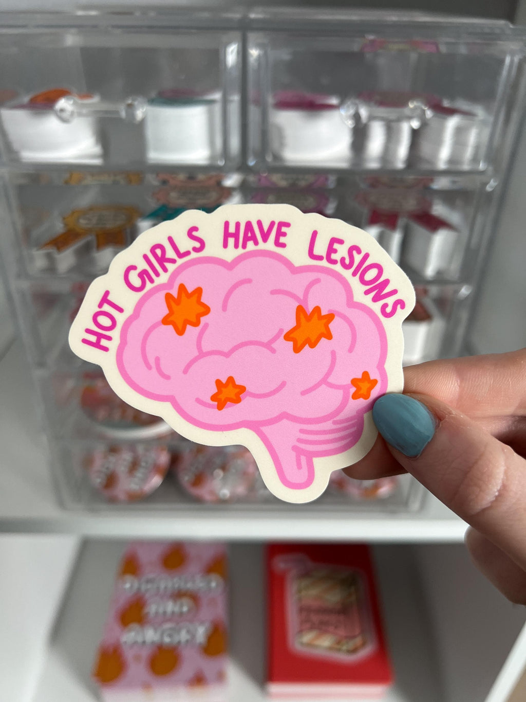 Brain shaped sticker with brain outlined in a darker bright pink and colored in with a light shade of bright pink. The brain has lesions on it that are orange. The text over the top of the brain in a hot pink reads, hot girls have lesions" with a cream background. The sticker is held in front of product display. 