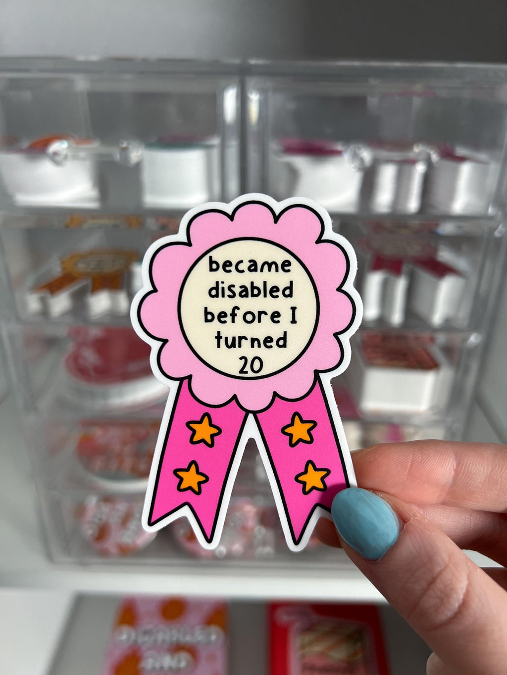 Award ribbon sticker in a light pink and dark pink shades, the middle is cream colored with the black font reading, "became disabled before I turned 20". Sticker is being held in front of product display. 