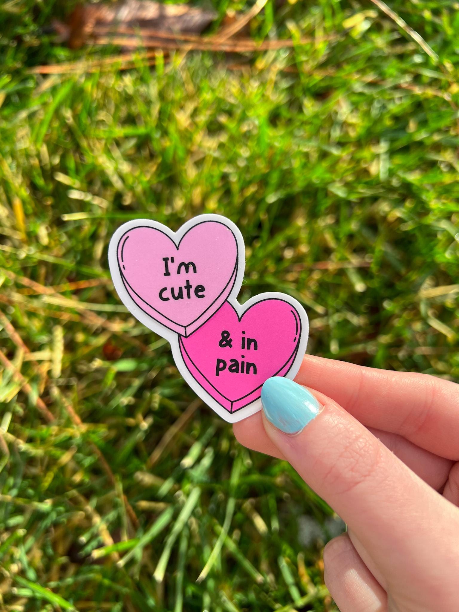 Candy hearts sticker. One heart in light pink and the other in a bright hot pink. The light pink heart reads, "I'm cute" while the hot pink reads, "and in pain". Sticker is held over grass. 